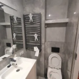 Szara łazienka z umywalką, toaletą i grzejnikiem na ręczniki z wieszakami w kształcie ludzików, widok z dołu.