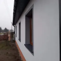 Świeżo otynkowana ściana zewnętrzna domu w Kutnie, z widocznymi oknami z szarymi obramowaniami i ciemną podbitką dachową, na tle pochmurnego krajobrazu.