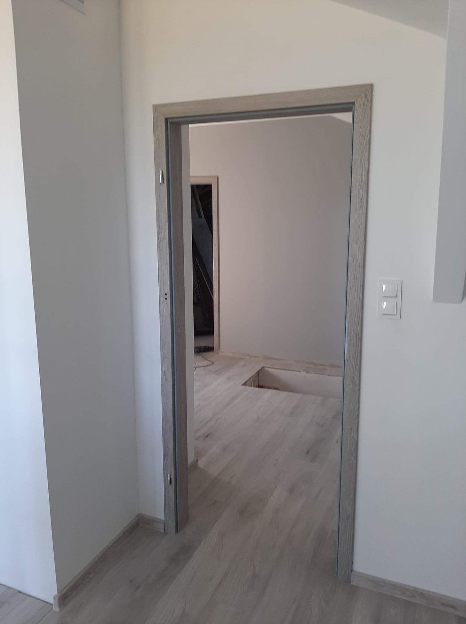 Otwór drzwiowy z szarą ościeżnicą w nowo wykończonym wnętrzu z panelami podłogowymi i włącznikami światła na ścianie. Widoczny otwór w podłodze w sąsiednim pomieszczeniu.
