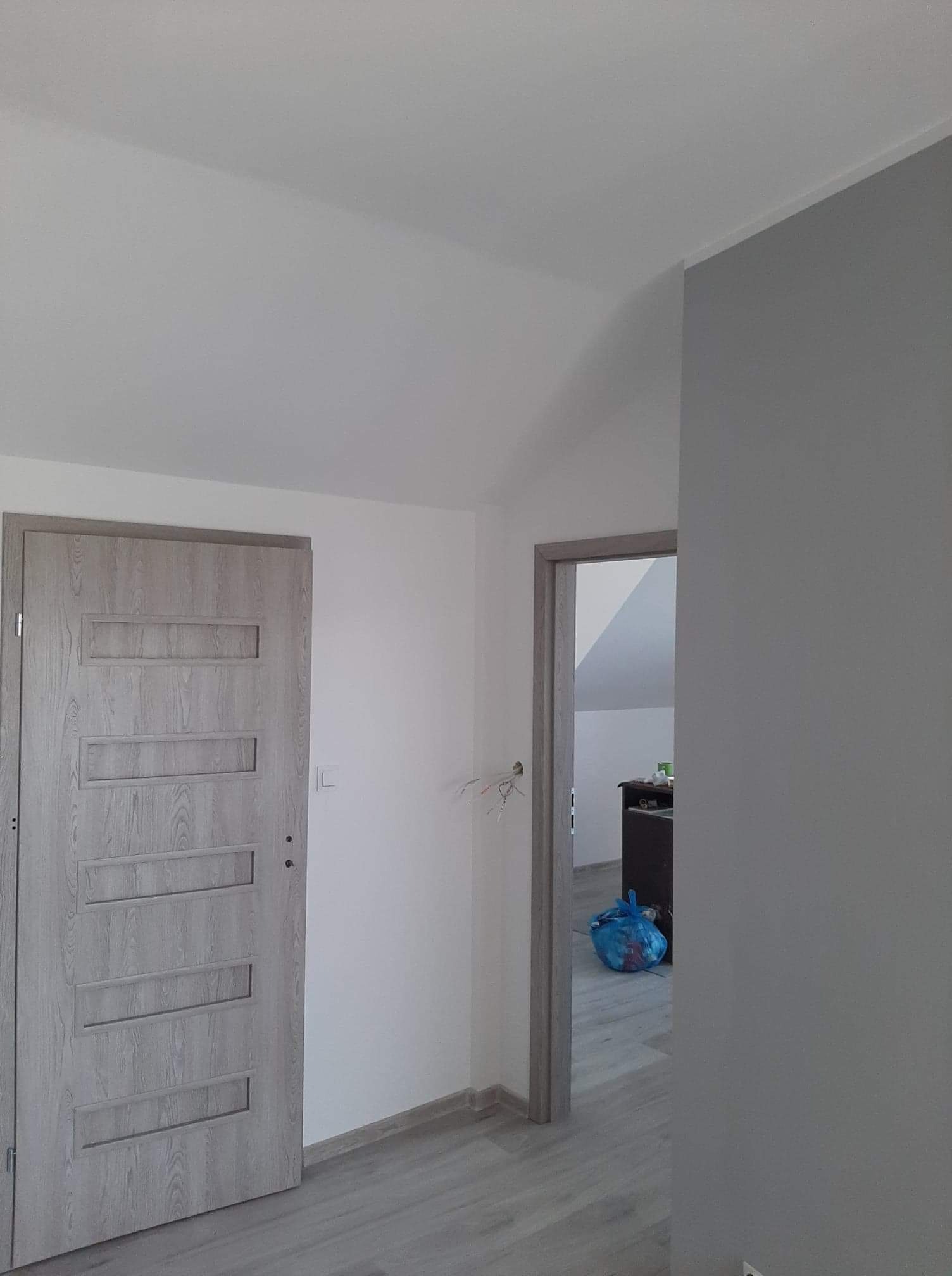 Wnętrze mieszkania po remoncie: szara ściana, drzwi wewnętrzne z drewnopodobnym wzorem i ościeżnicą w tym samym kolorze, widok na pokój z workiem na śmieci.