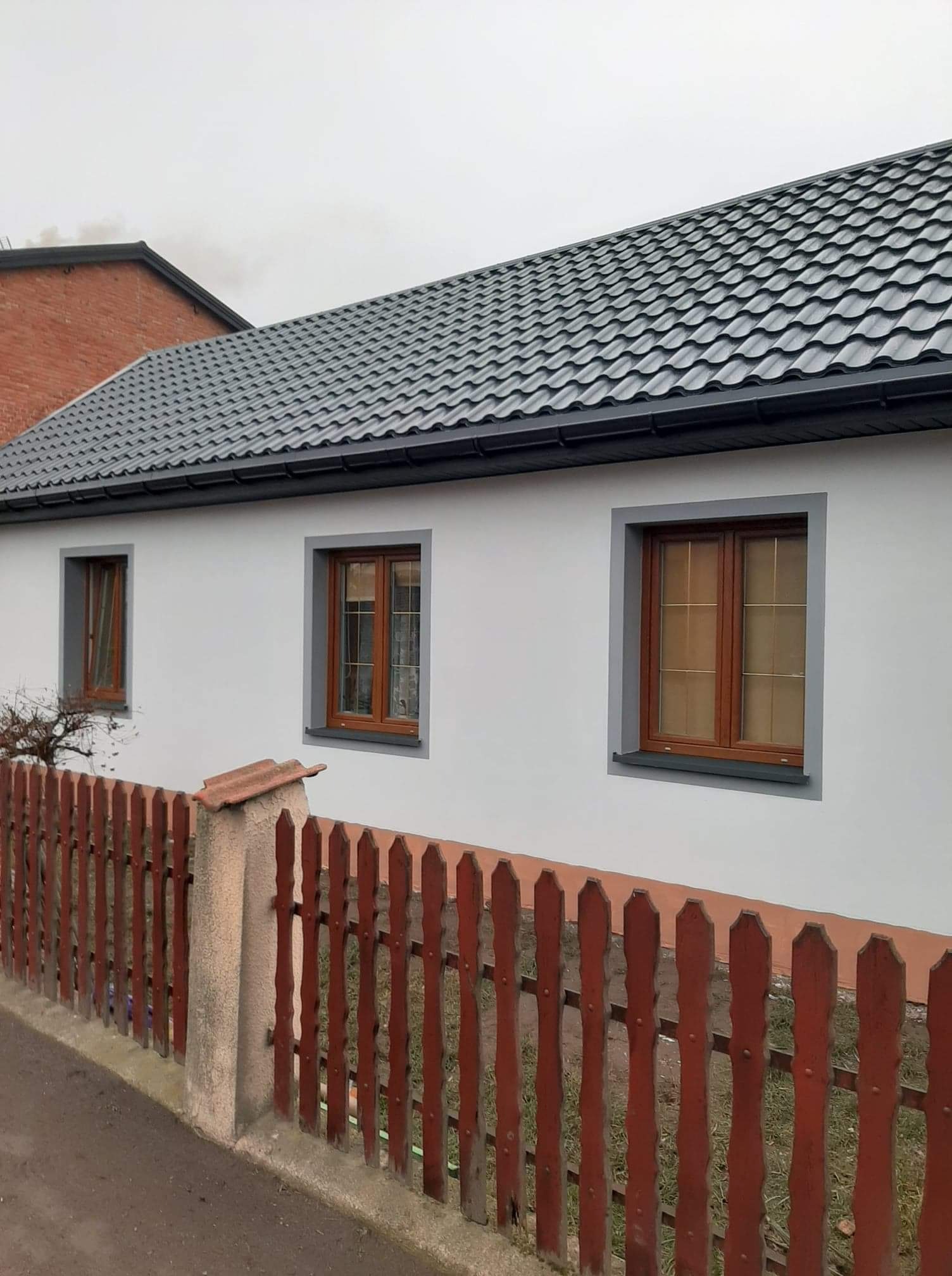 Świeżo otynkowany dom jednorodzinny z szarą dachówką i brązowymi oknami, widziany zza niskiego, czerwonego płotu.