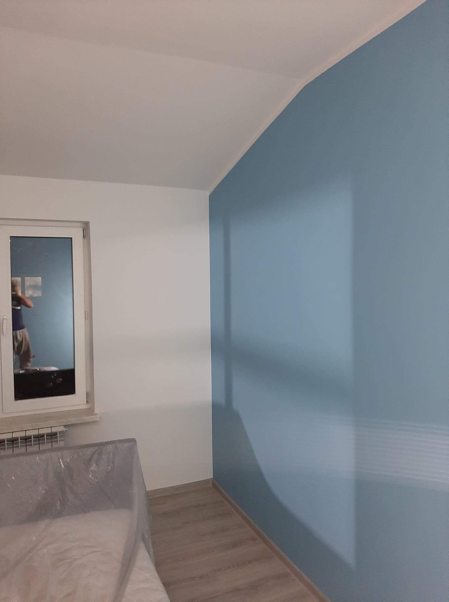 Świeżo pomalowane wnętrze: błękitna ściana kontrastuje z białą, okno z grzejnikiem, meble zabezpieczone folią malarską, parkiet na podłodze.