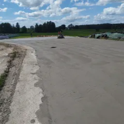 Świeżo wylana, szara, betonowa posadzka przemysłowa z widocznymi śladami po zacieraczce mechaniczną, w tle zielone pola i błękitne niebo z białymi chmurami.