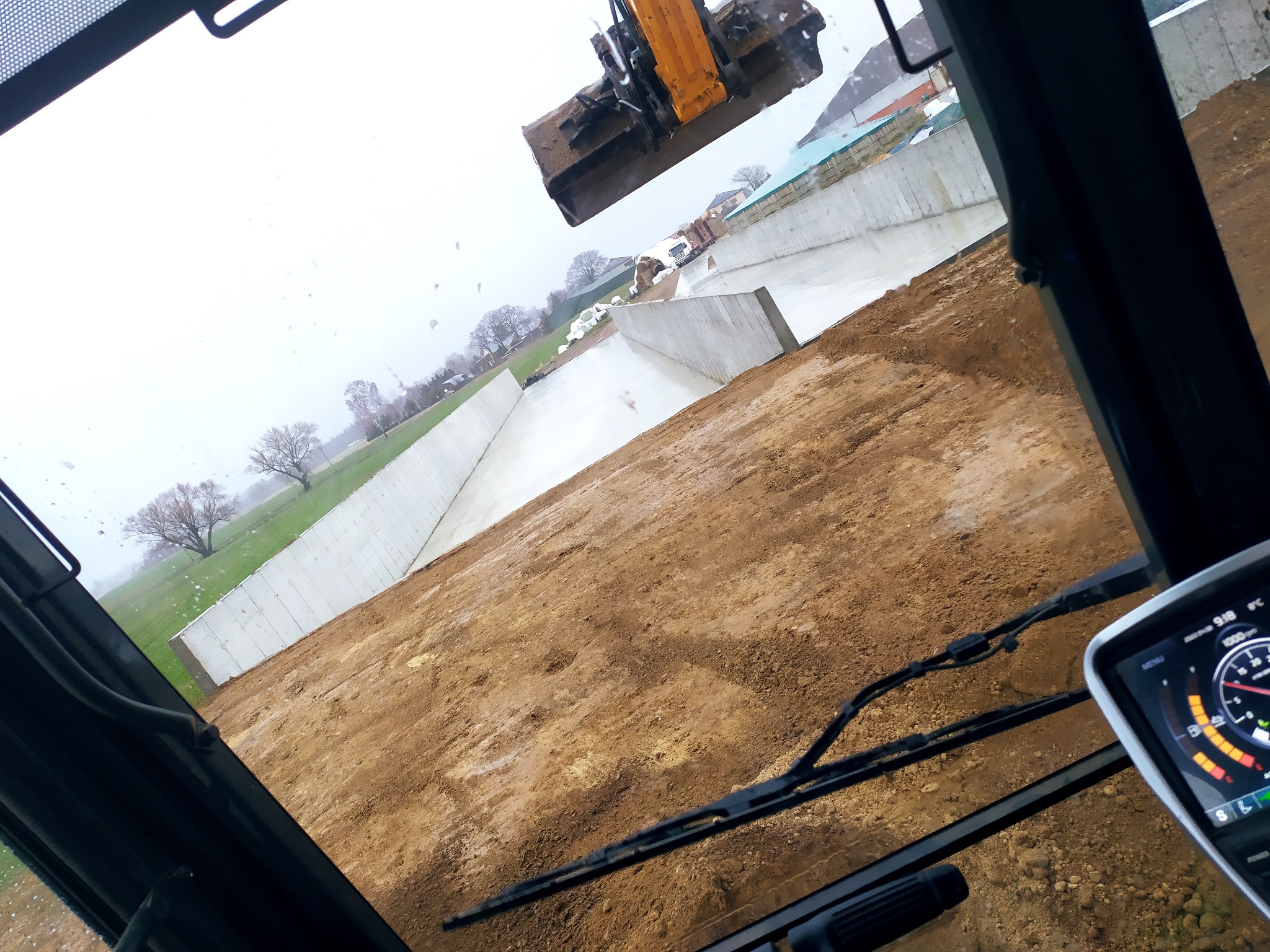 Widok z kabiny koparki na plac budowy z wykopem ograniczonym betonowymi ścianami, na zewnątrz zielone łąki i drzewa w pochmurny dzień. Wykoparka nad wykopem.