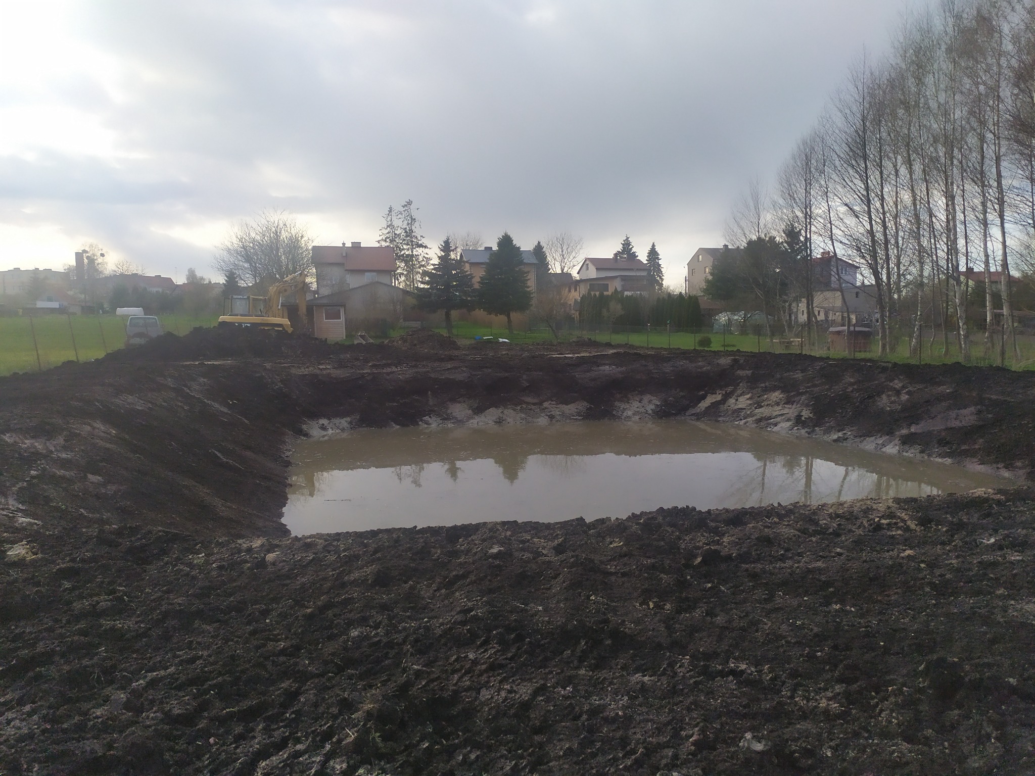 Wykopany staw wypełniony wodą, brzeg z ciemnej ziemi, w tle zabudowania i drzewa, widoczna koparka na skraju wykopu.