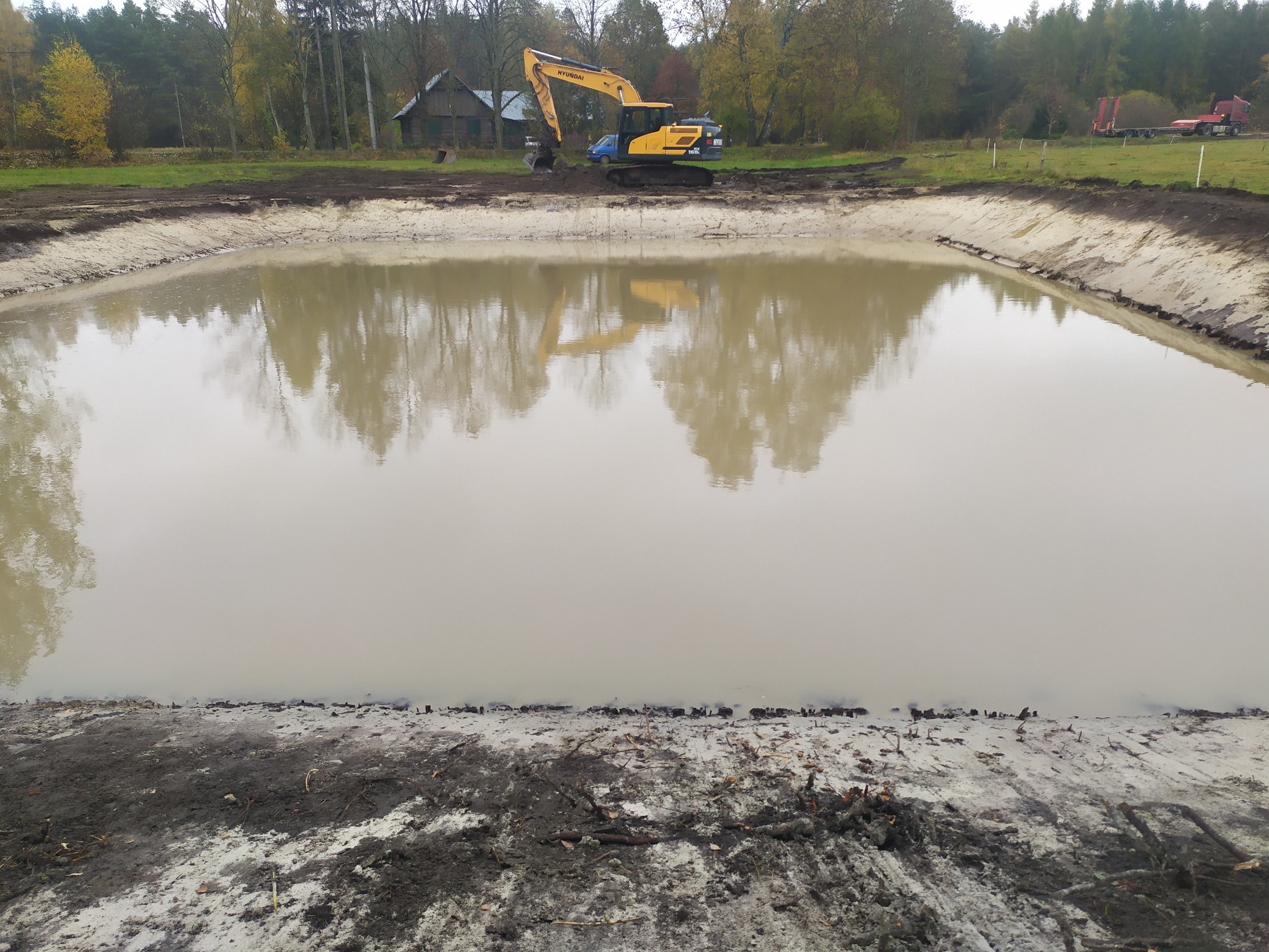 Żółto-niebieska koparka Hyundai na brzegu nowo wykopanego stawu, którego powierzchnia wody odbija drzewa i niebo; w tle zabudowania gospodarcze i drzewa.