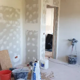 Wnętrze w trakcie prac wykończeniowych: ściany i sufit pokryte warstwą gipsową, widoczne narzędzia i materiały budowlane, w tle otwarte przejście do innego pomieszczenia.