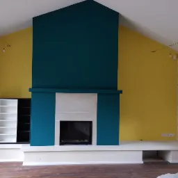 Wnętrze pokoju z kominkiem, ściany pomalowane na żółto i turkusowo, jedna ściana tapetowana w delikatny wzór, na suficie szynoprzewód z reflektorami, drewniana podłoga.