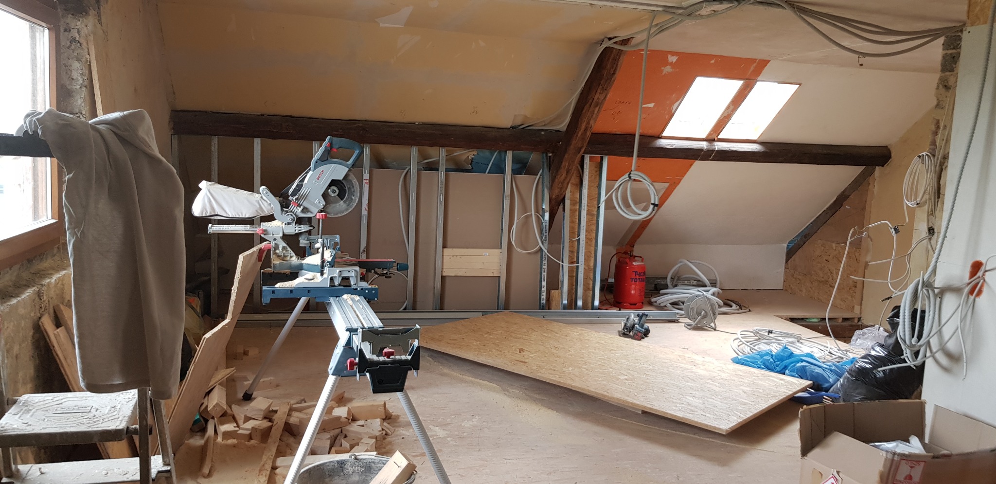 Adaptacja poddasza: widok na wnętrze w trakcie prac, z widoczną konstrukcją ścian działowych, nowymi oknami dachowymi, przewodami elektrycznymi oraz narzędziami, w tym piłą ukośnicą marki Bosch...