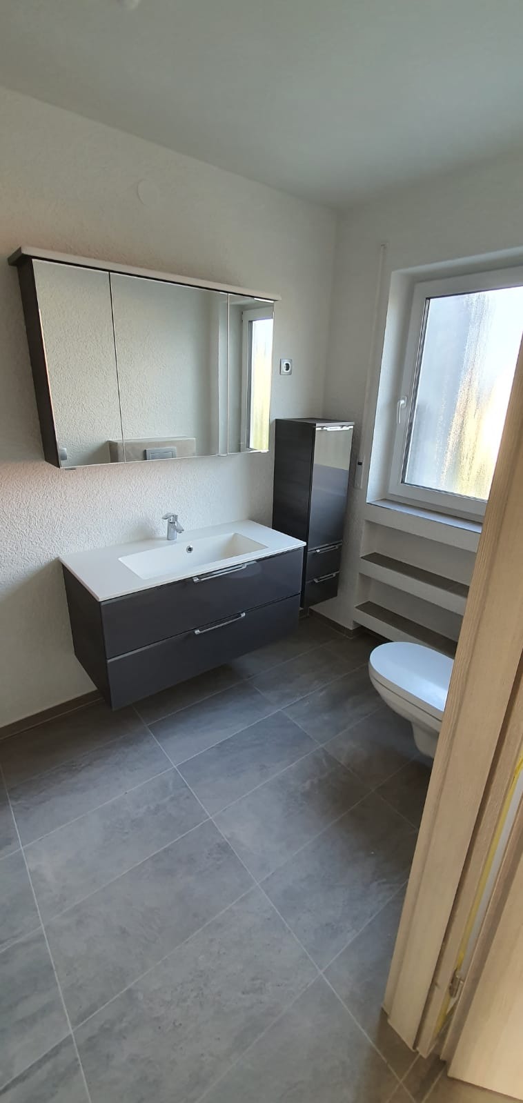 Nowoczesna łazienka z szarymi płytkami podłogowymi, wiszącą szafką z umywalką, lustrem z szafkami i wysoką szafką obok toalety przy oknie.