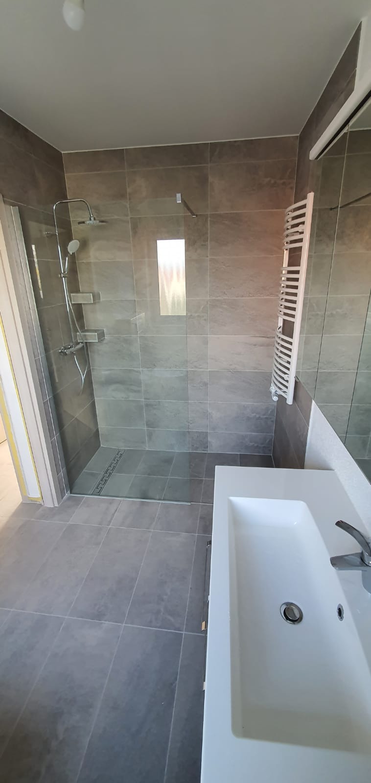 Wykończona łazienka z szarymi płytkami, kabiną prysznicową typu walk-in z odpływem liniowym, grzejnikiem drabinkowym i białą umywalką.