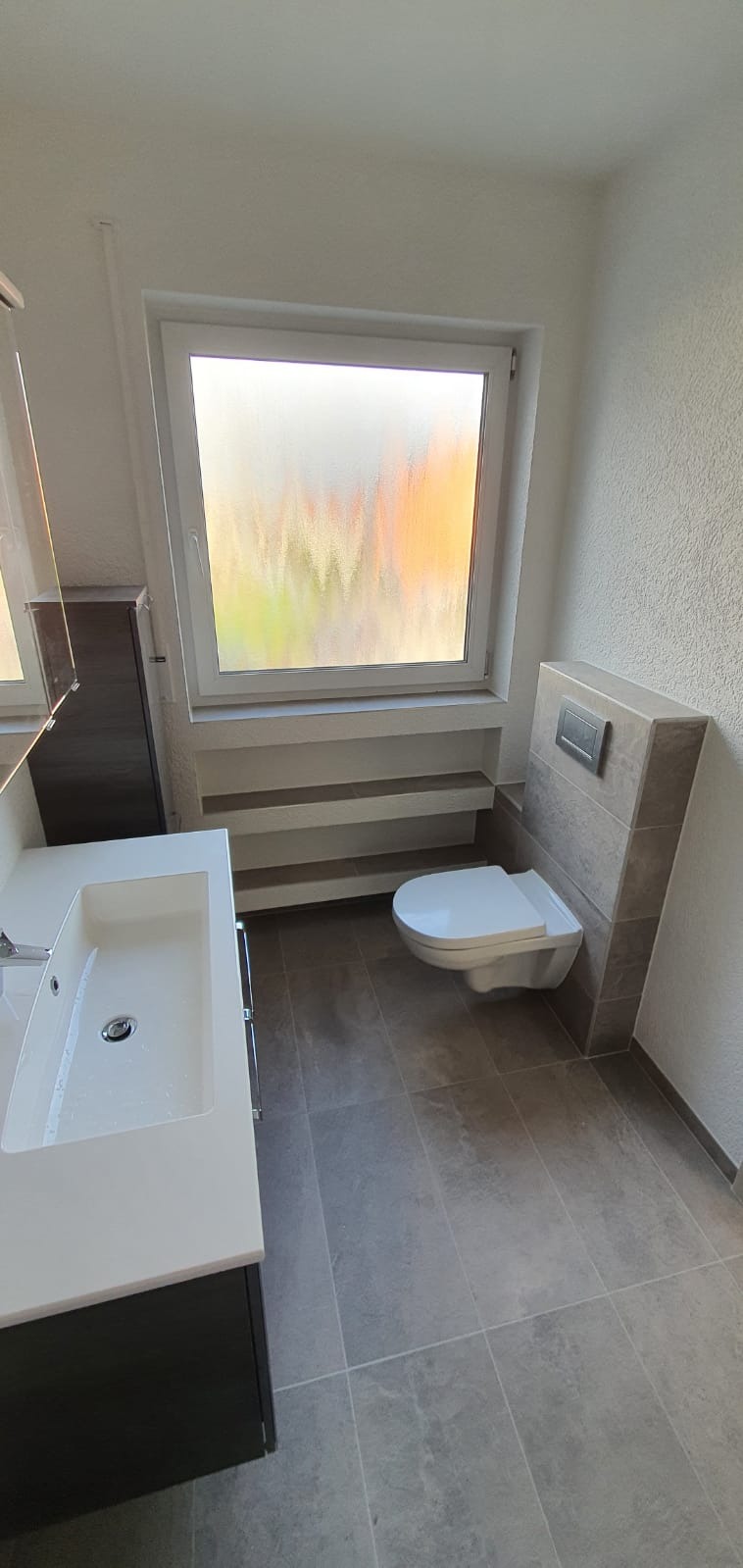 Nowoczesna łazienka z podwieszaną toaletą, umywalką z szafką w kolorze antracytowym i szarą glazurą podłogową oraz oknem z szybą ornamentową.