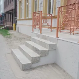 Nowe, jasnoszare, kamienne schody wejściowe do budynku z ozdobną, metalową balustradą w kolorze miedzianym.