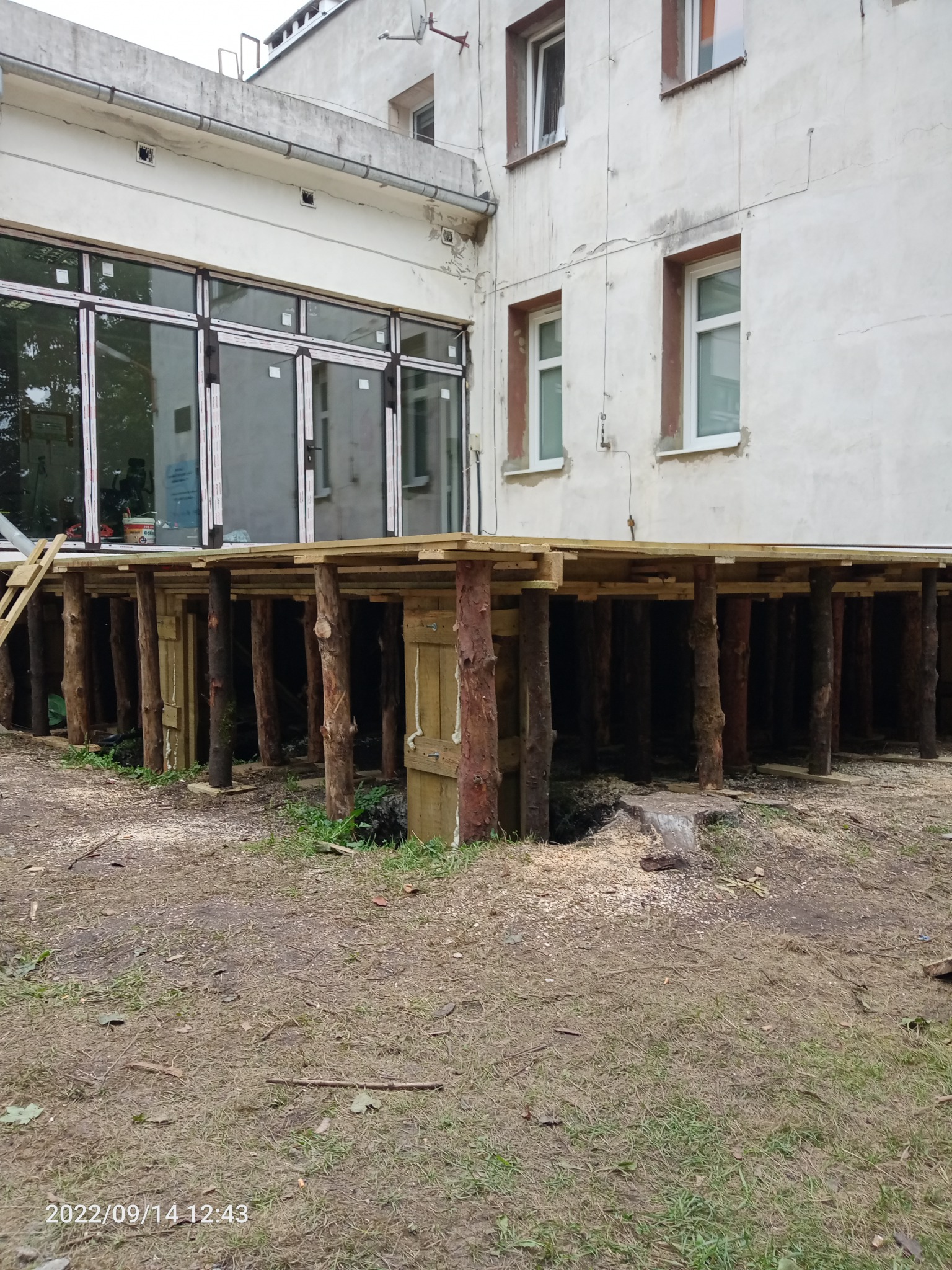 Wzmocnienie fundamentów budynku z użyciem drewnianych bali i belek, widoczne ślady wykopów i obróbki drewna, budynek z oknami w tle.