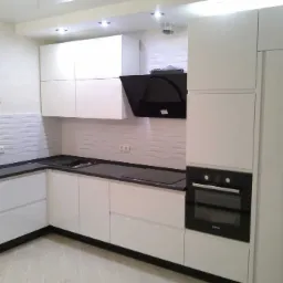 Nowoczesna biała kuchnia z czarnymi blatami, minimalistyczna zabudowa z piekarnikiem, czarny okap i lodówka, panel ścienny z efektem falowania, drewnopodobna podłoga.