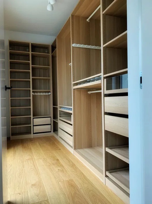 Jasna, drewniana garderoba z półkami, szufladami i relingami na ubrania, widok z perspektywy wejścia, podłoga z paneli, oświetlenie sufitowe.
