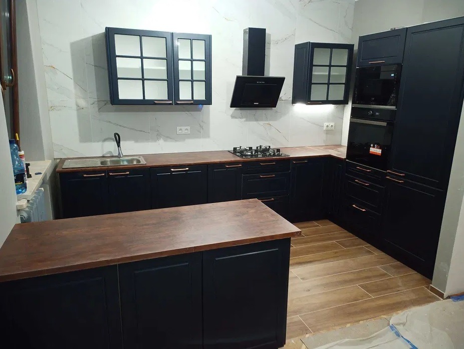 Nowoczesna kuchnia w ciemnej kolorystyce z drewnianymi blatami, widok na zabudowę kuchenną z wyspą, płytą grzewczą i piekarnikiem, marmurowe ściany w tle.