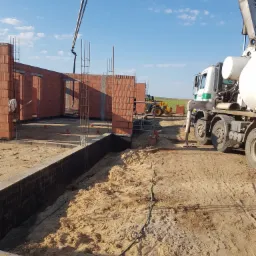 Wylewanie betonu do fundamentów domu z częściowo wymurowanymi ścianami z czerwonej cegły, widoczny betonowóz i pompa do betonu na tle błękitnego nieba.