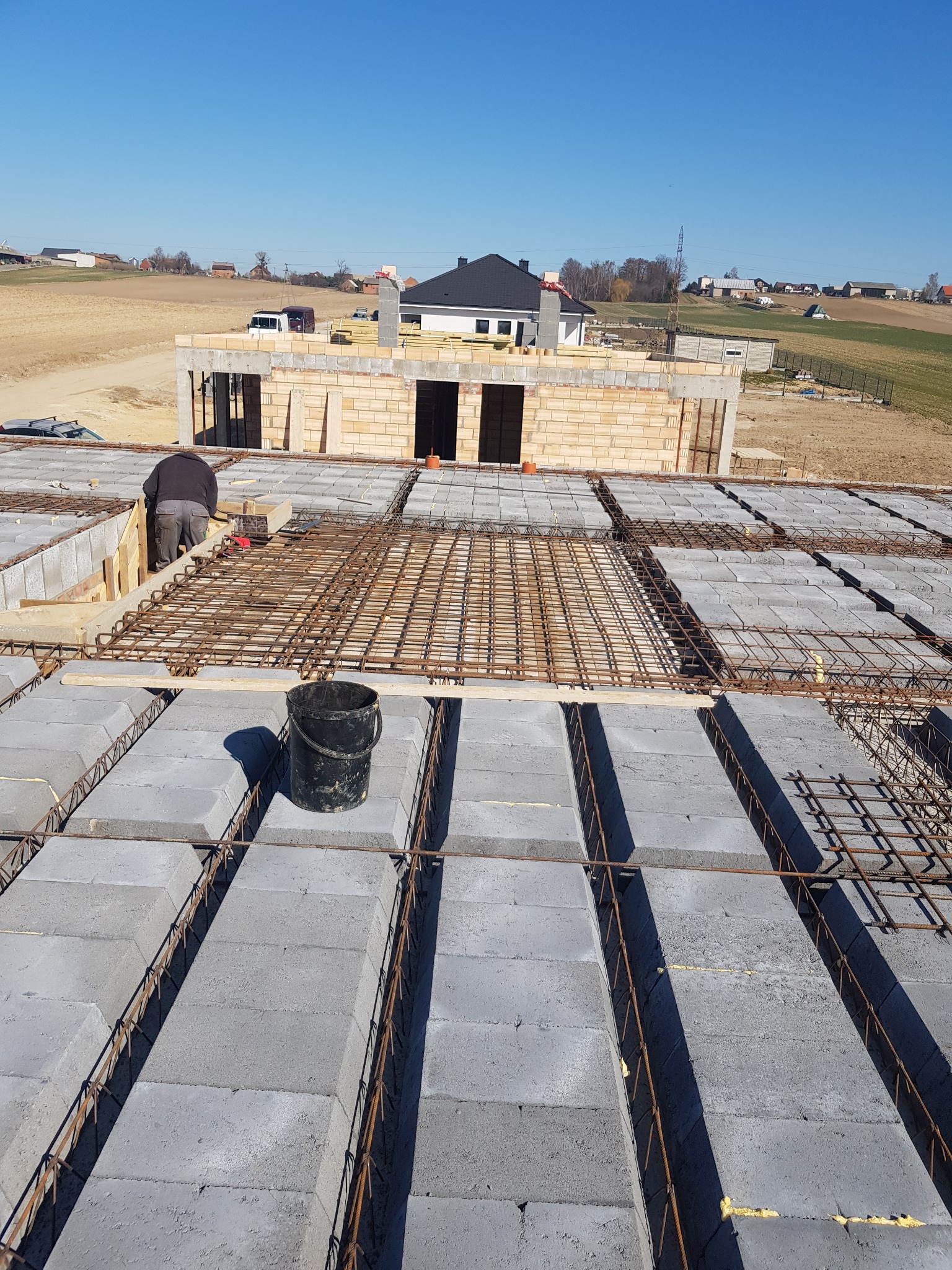 Budowa domu: widok z góry na strop Teriva z ułożonym zbrojeniem, w tle wzniesione ściany parteru i krajobraz wiejski.