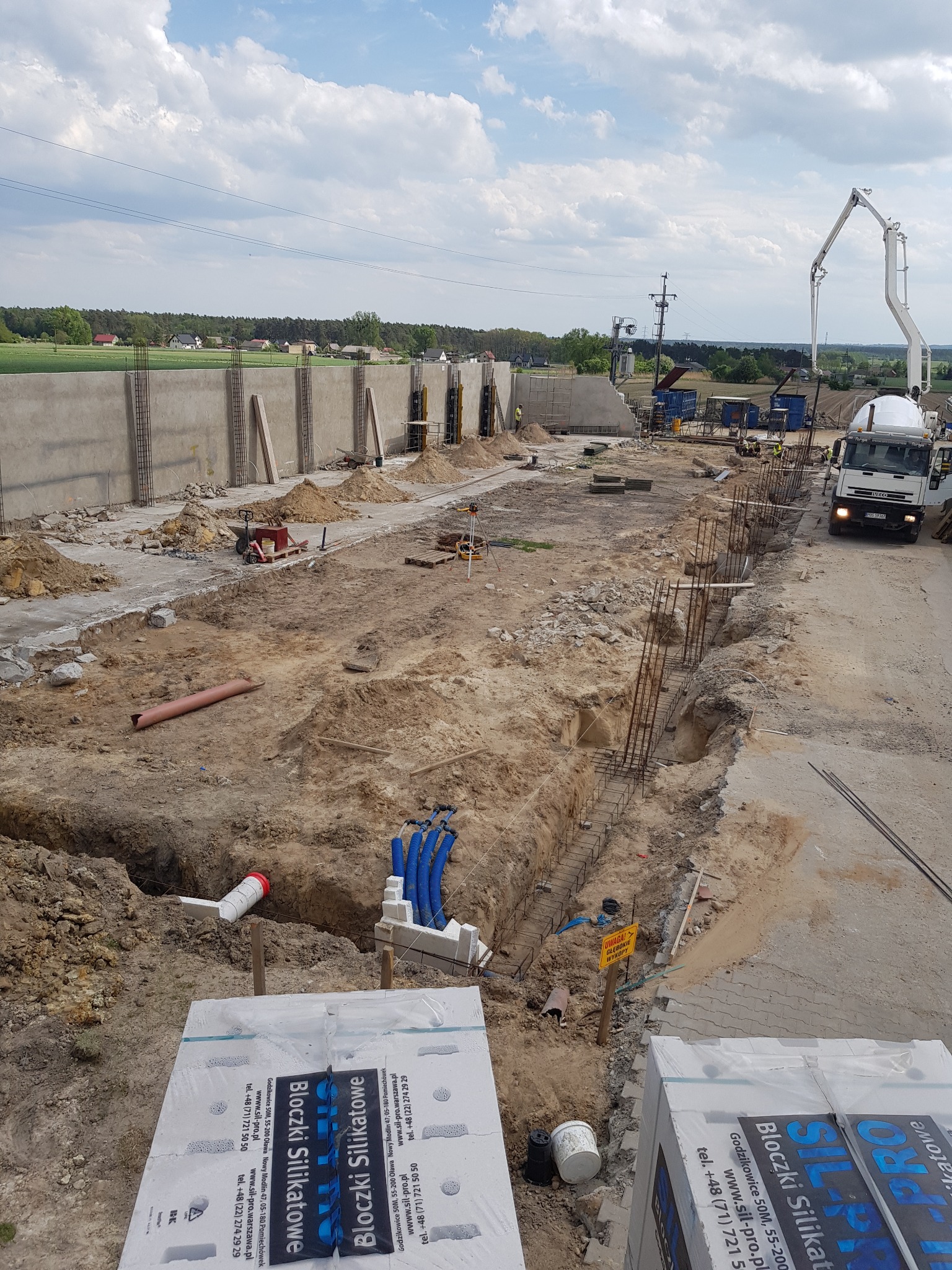 Budowa fundamentów: widok na wykop zbrojony stalą, z betonową pompą w tle oraz paletami bloczków silikatowych na pierwszym planie.