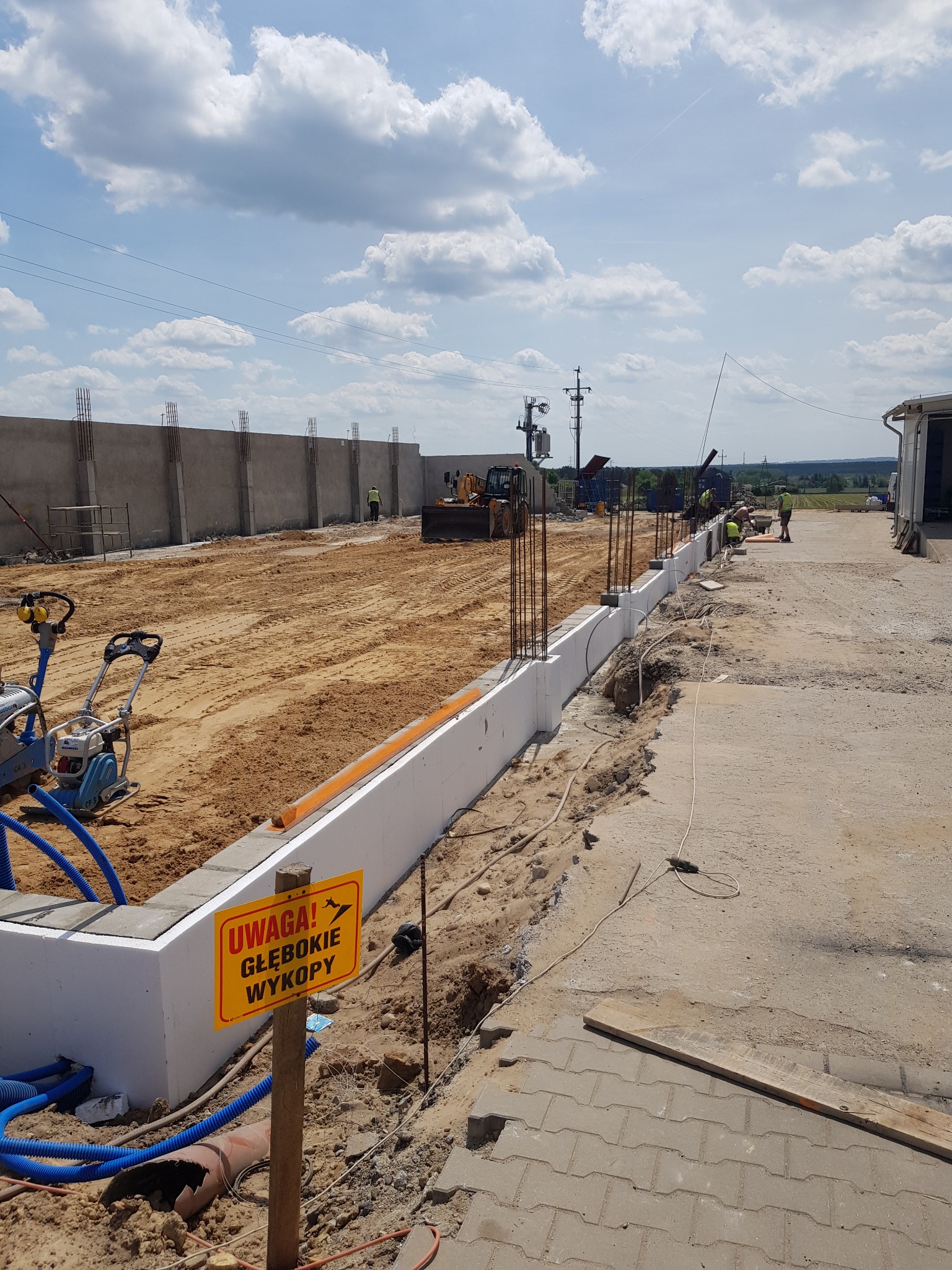 Budowa fundamentów: widok na wykop z częściowo wylanym betonem, wystającymi prętami zbrojeniowymi, żółtą tablicą ostrzegawczą 'Uwaga! Głębokie Wykopy' i sprzętem budowlanym w tle.