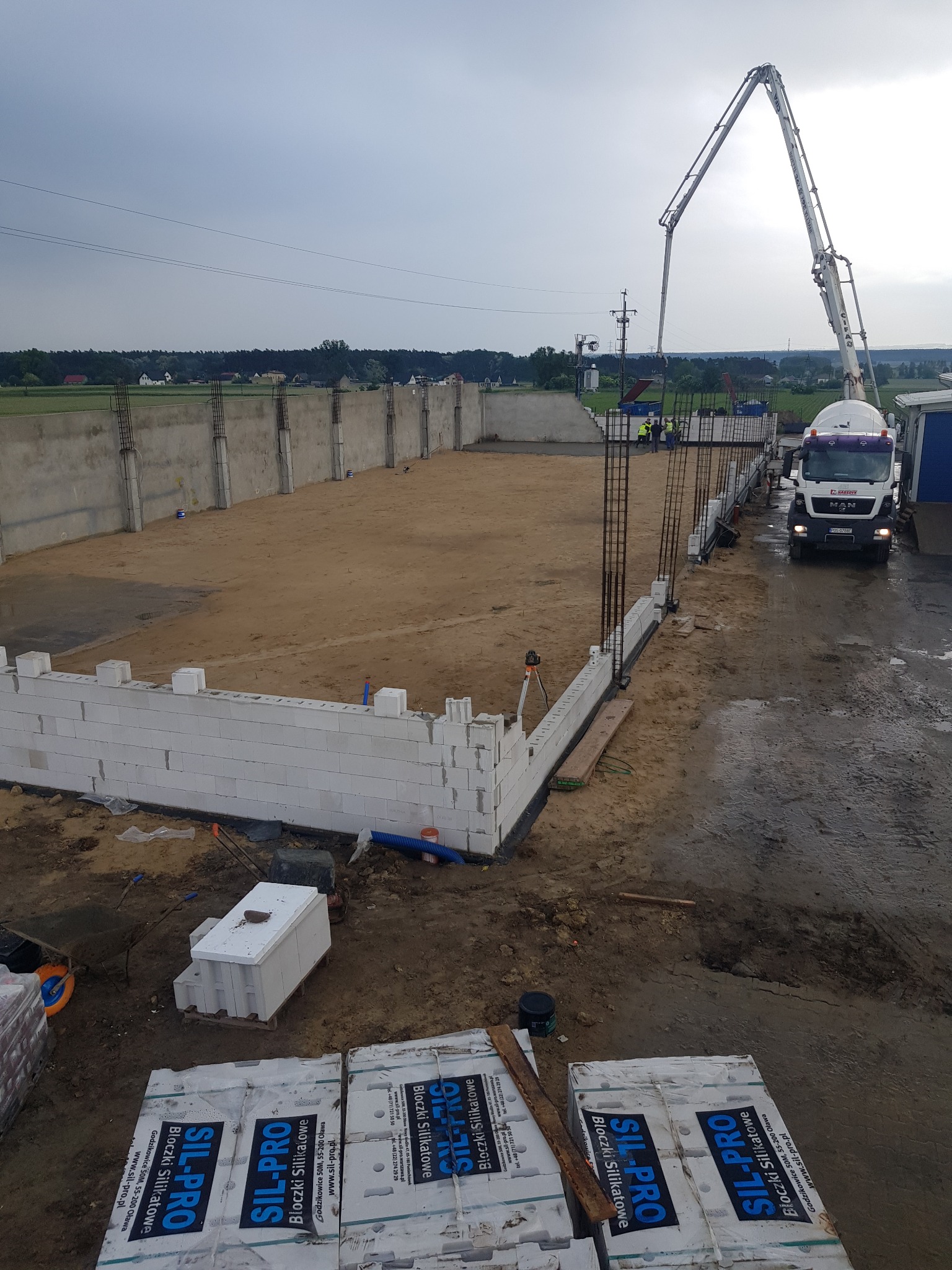 Budowa domu z bloczków silikatowych Sil-Pro, widoczny betonowóz z pompą, zbrojenia i fragmenty ścian fundamentowych na placu budowy w pochmurny dzień.