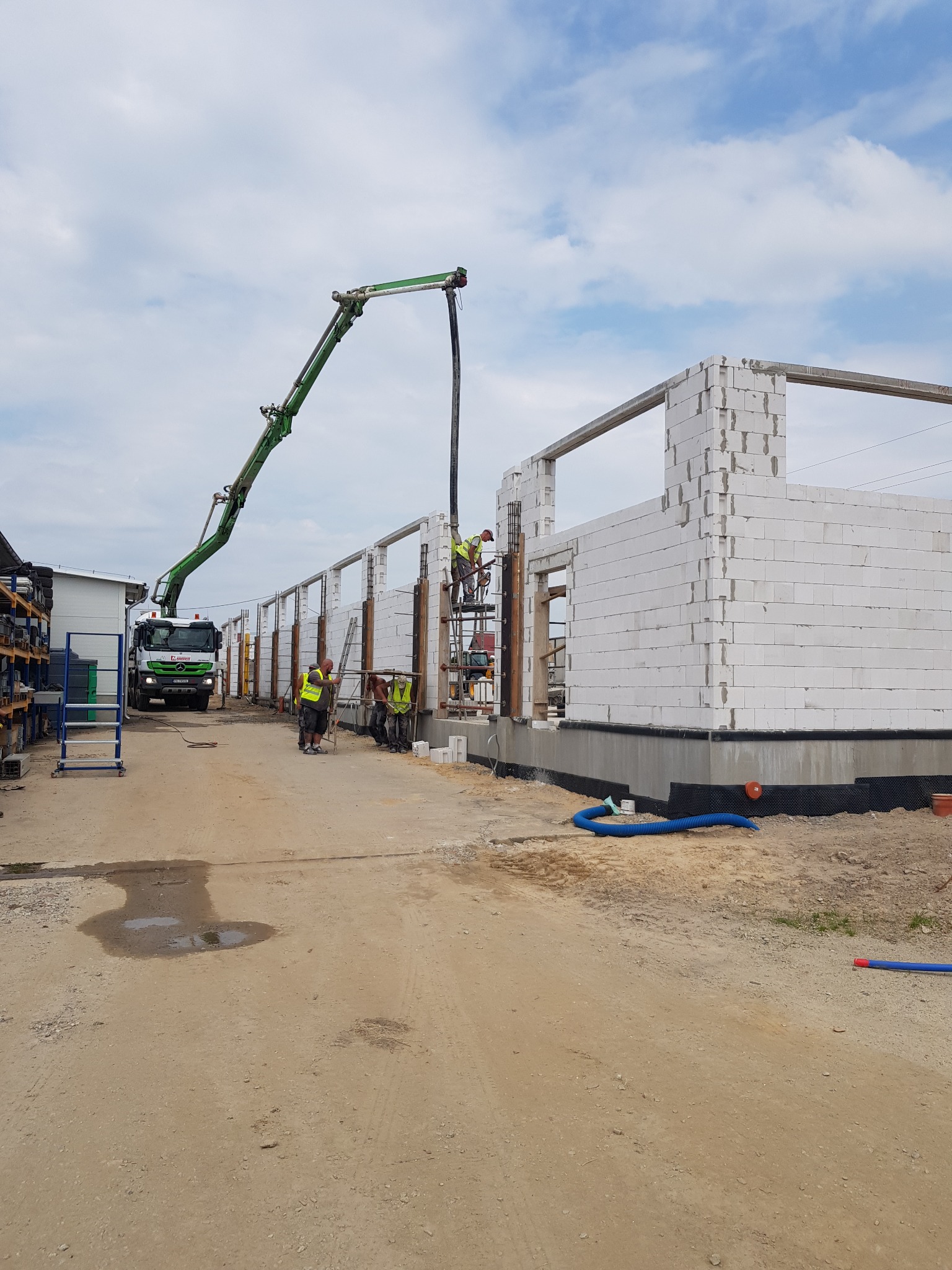 Wznoszenie ścian budynku z betonu komórkowego; widoczna pompa do betonu, pracownicy na rusztowaniach i fragmenty ścian z otworami okiennymi.