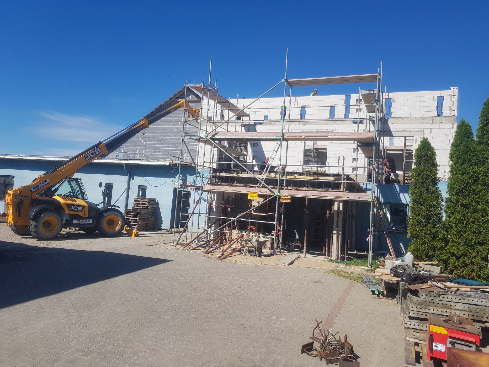 Wznoszenie dwukondygnacyjnego budynku z białych bloczków, widoczne rusztowania, podnośnik teleskopowy JCB i pracownicy na budowie w słoneczny dzień.