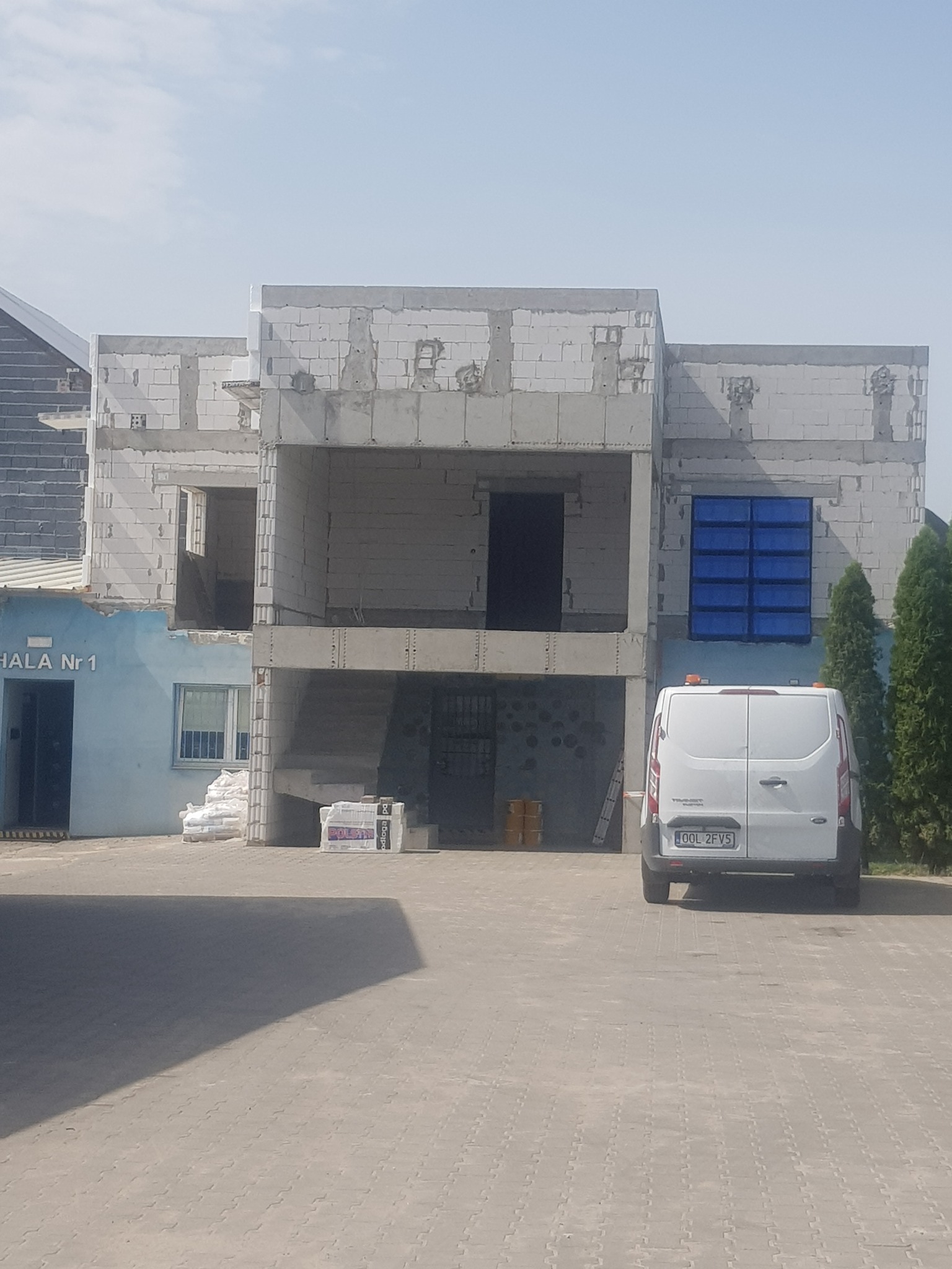 Stan surowy otwarty dwukondygnacyjnego budynku z białego bloczka w Czekanowie, widoczny z zewnątrz, z zaparkowanym białym busem i stertą materiałów budowlanych.