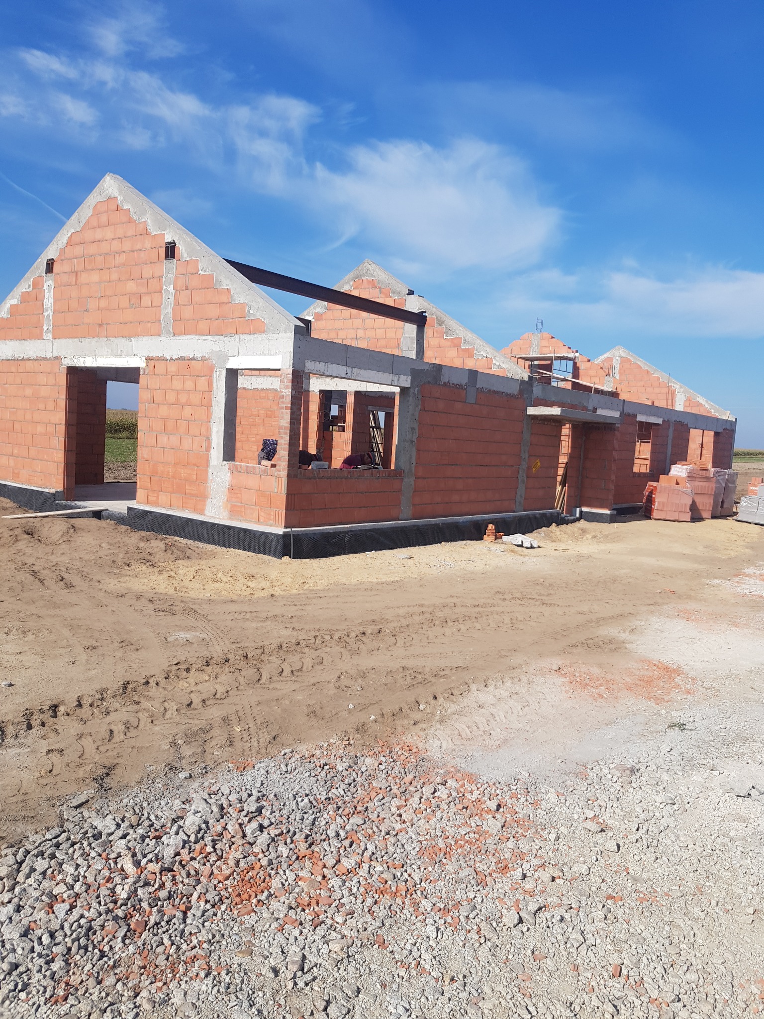 Wznoszenie ścian z czerwonej cegły ceramicznej na budowie domu jednorodzinnego, widoczne okna i drzwi bez stolarki, nieotynkowane ściany szczytowe, podłoże z gruzu i piasku.