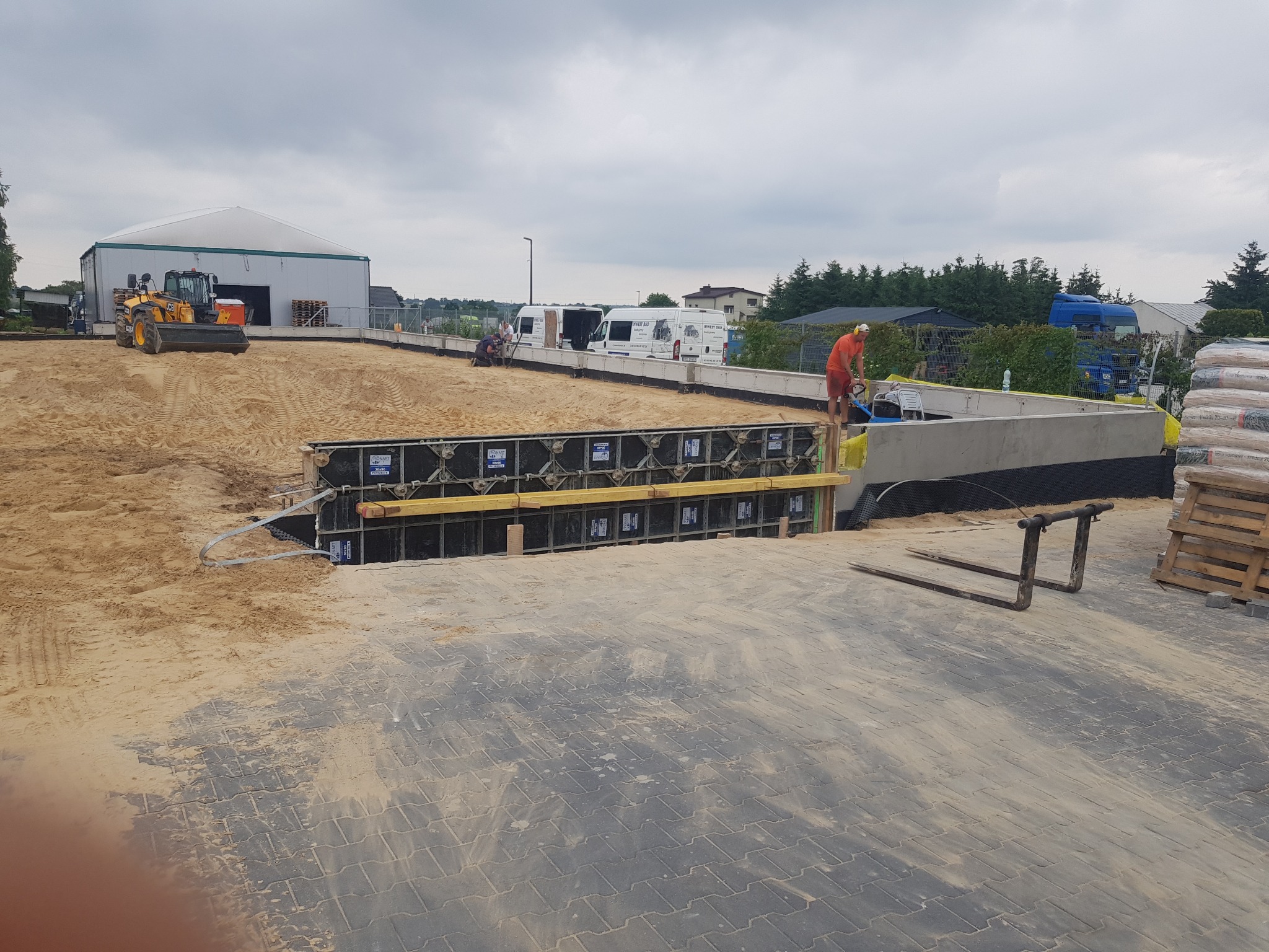 Budowa fundamentów: pracownik w pomarańczowym stroju przycina deskę, widoczne szalunki, wykop, maszyna budowlana w tle, teren przygotowany pod wylewkę.