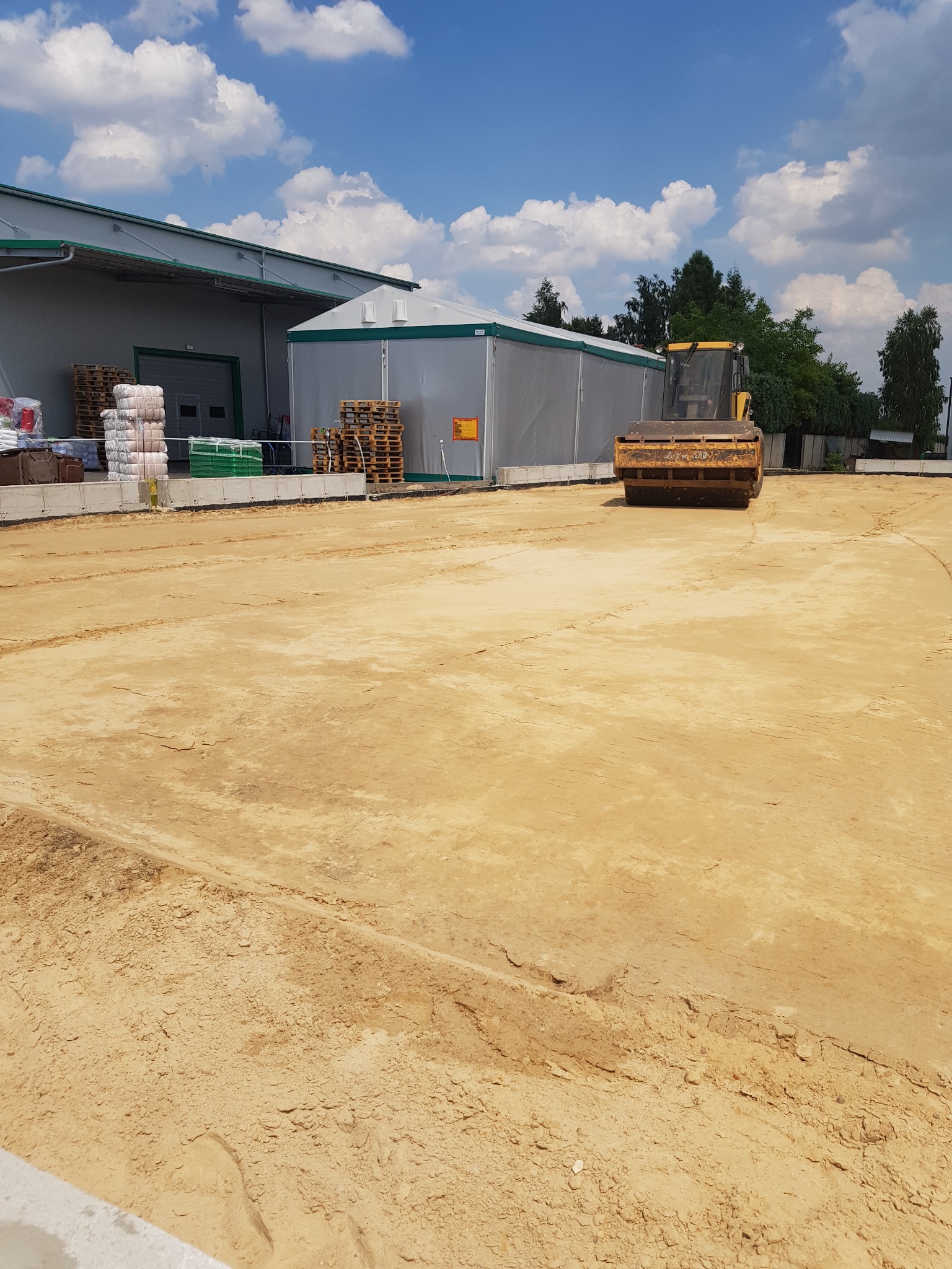 Żółty walec drogowy ubija piasek na placu budowy, widoczne magazyny i materiały budowlane w tle, błękitne niebo z chmurami.