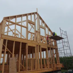 Konstrukcja szkieletowa domu w trakcie budowy, widoczni dwaj pracownicy na rusztowaniu, drewniane elementy konstrukcyjne, pochmurne niebo w tle.