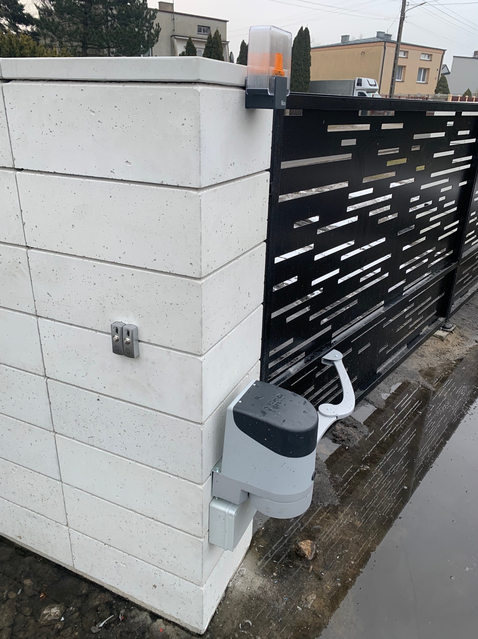 Nowoczesna, czarna brama z automatyką NICE zamontowana na białym, betonowym słupku. Widoczny napęd i lampa ostrzegawcza. Błoto na ziemi.