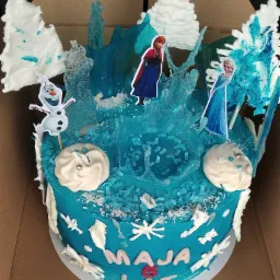 Tort urodzinowy w stylu Krainy Lodu, z imieniem 'Maja 5 lat', ozdobiony postaciami Elsy, Anny i Olafa, oraz lukrowanymi drzewkami i lodowymi soplami, na niebieskim tle z posypką.