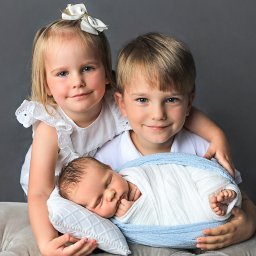 ANGEL WINGS PHOTOGRAPHY Natalia Desyatova - Uśmiechnięta dziewczynka z kokardą i chłopiec obejmujący śpiącego noworodka owiniętego w biało-niebieski kocyk, leżącego na poduszce na szarym tle.