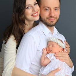 ANGEL WINGS PHOTOGRAPHY Natalia Desyatova - Portret szczęśliwej rodziny: uśmiechnięta matka obejmuje ojca trzymającego śpiące nowonarodzone dziecko ubrane w białą koronkową sukienkę i wianek na głowie, na jednolitym, ciemnoszarym tle.
