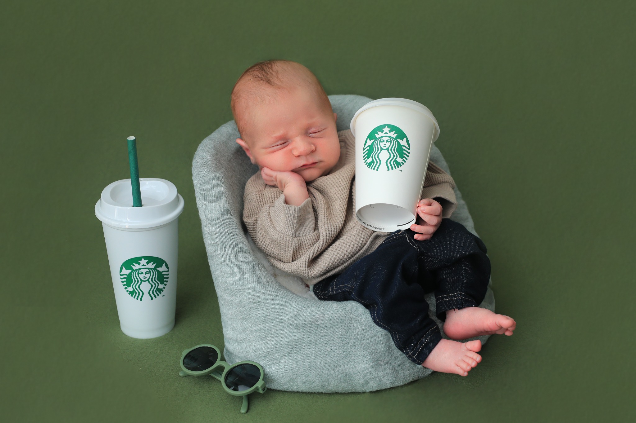 Noworodek śpiący w foteliku, trzymający kubek Starbucks, obok drugi kubek z zieloną słomką i małe zielone okulary na zielonym tle.