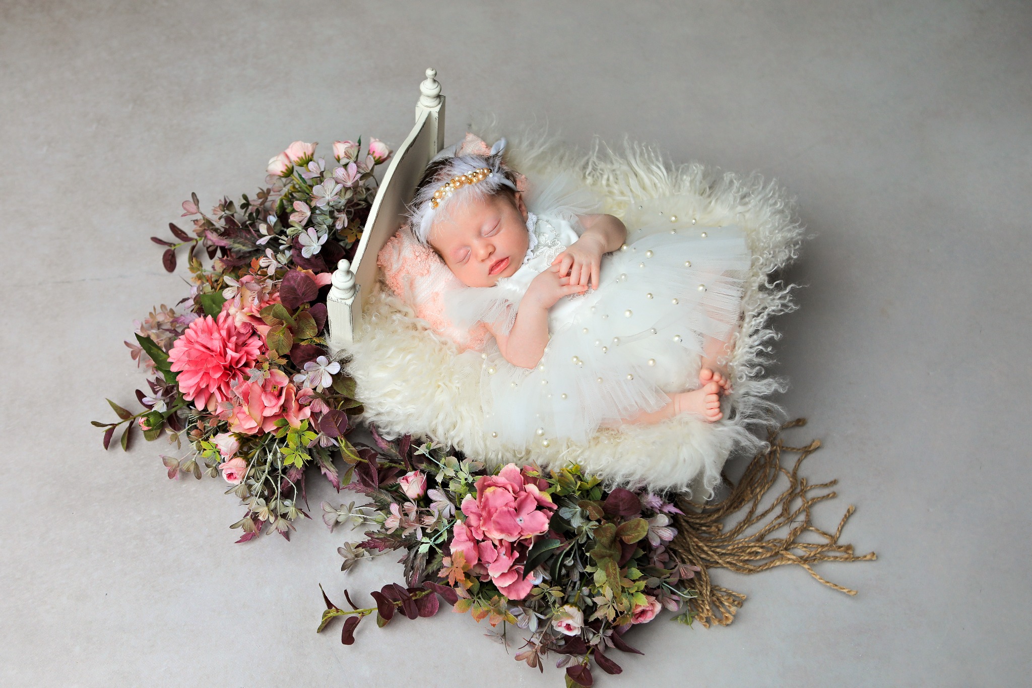 Śpiąca noworodka w białej sukience z perełkami, leżąca w małym łóżeczku otoczonym kwiatami i futerkiem na szarym tle.