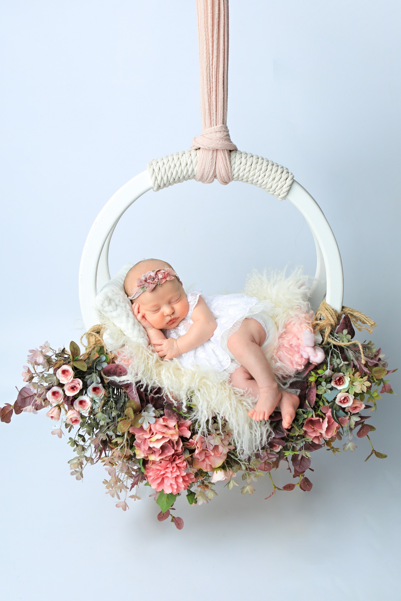 Śpiąca noworodka w białej sukience i opasce z kwiatami, ułożona w okrągłym, białym koszu zawieszonym na różowej tkaninie, otoczonym dekoracjami z różowych kwiatów i futerkiem.