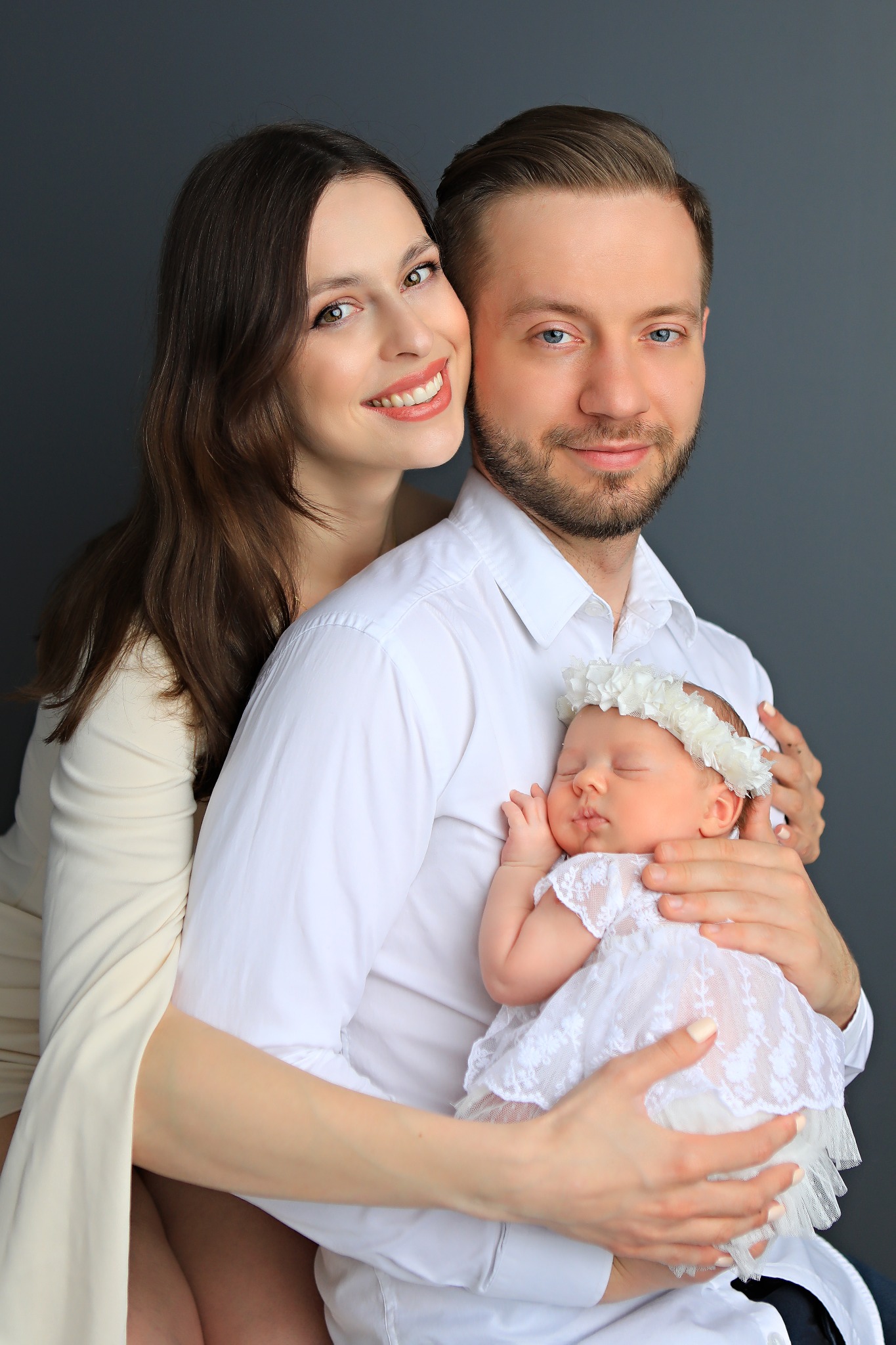 Portret szczęśliwej rodziny: uśmiechnięta matka obejmuje ojca trzymającego śpiące nowonarodzone dziecko ubrane w białą koronkową sukienkę i wianek na głowie, na jednolitym, ciemnoszarym tle.