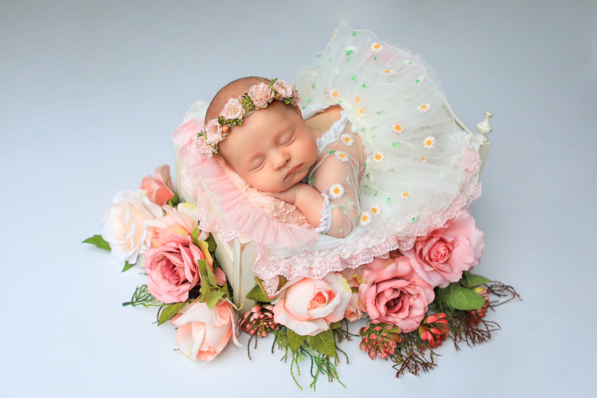Noworodek śpiący w małym łóżeczku otoczonym różowymi i białymi różami, ubrany w białą sukienkę w stokrotki i wianek z różowych kwiatów na głowie.
