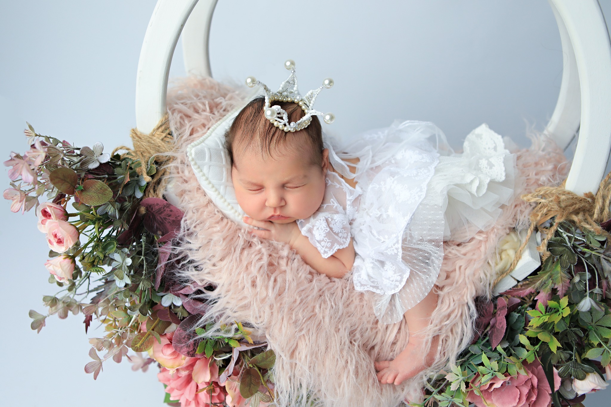 Śpiąca noworodka w koronie na poduszce z jasnoróżowego futerka, w białej koronkowej sukience, otoczona kwiatami w okrągłym, białym koszu.