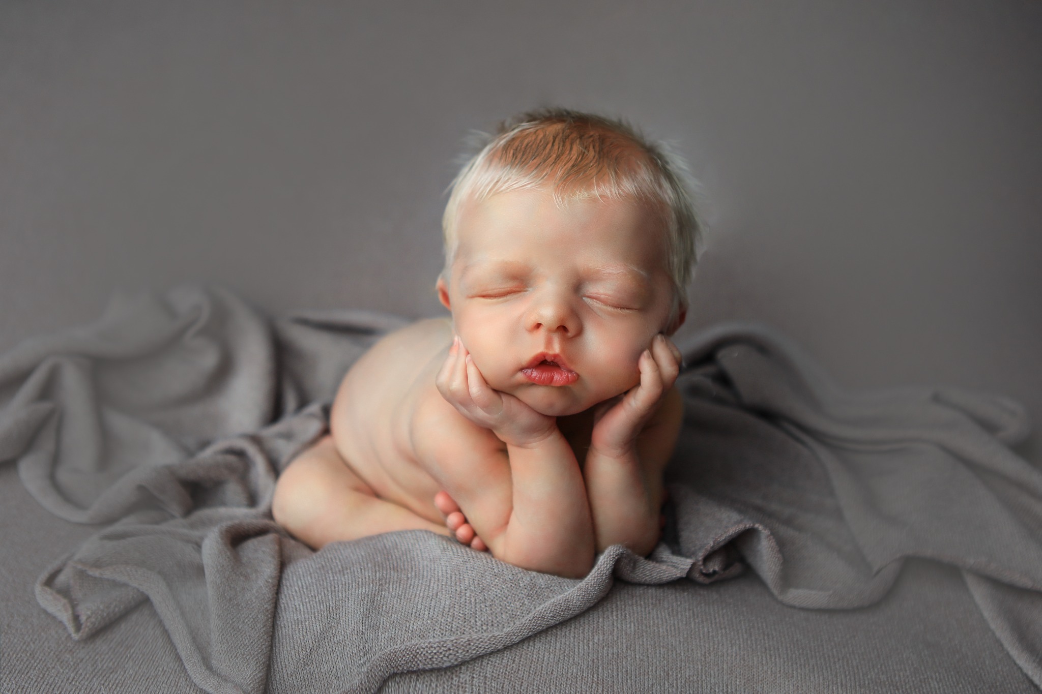 Śpiący noworodek z jasnymi włosami, podpierający buzię rączkami, ułożony na szarym, miękkim kocyku.