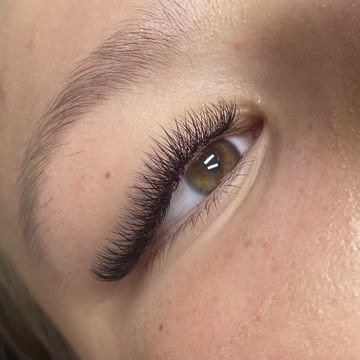 Zbliżenie na oko z wyraźnie wydłużonymi i zagęszczonymi rzęsami, widoczne precyzyjne połączenia sztucznych rzęs z naturalnymi, brwi starannie wystylizowane.