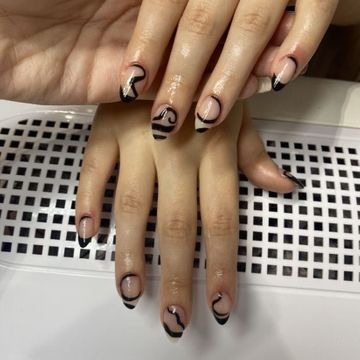 Dłonie z manicure hybrydowym: naturalna płytka, czarne abstrakcyjne wzory, cienkie czarne końcówki na niektórych paznokciach, widoczne delikatne refleksy światła.