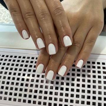 Dłonie z białym, jednolitym lakierem na krótkich, kwadratowych paznokciach, ułożone na kratownicy. Widoczne delikatne refleksy światła na powierzchni lakieru.