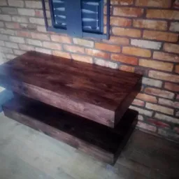 Dwupoziomowa, ciemnobrązowa półka z litego drewna, zamontowana na tle ściany z cegły w odcieniach pomarańczy i beżu, oświetlona z góry.