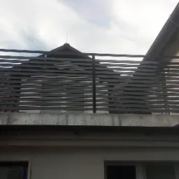 Nowoczesna, pozioma, metalowa balustrada balkonowa w kolorze antracytowym, zamontowana na betonowej płycie, widok z dołu na tle nieba i dachu budynku.