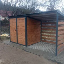 Drewniany schowek na narzędzia z zadaszeniem, obok ułożone drewno na tle domu w budowie i drzew bez liści.